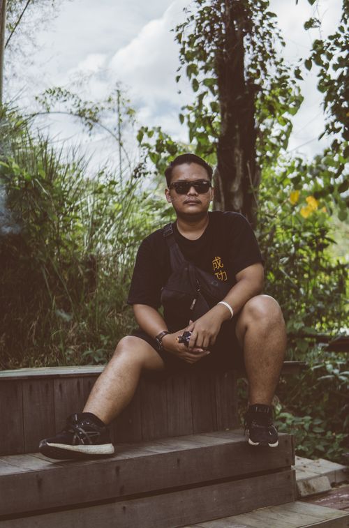Free A man in a black shirt and sunglasses poses sitting on wooden steps surrounded by lush greenery. Stock Photo