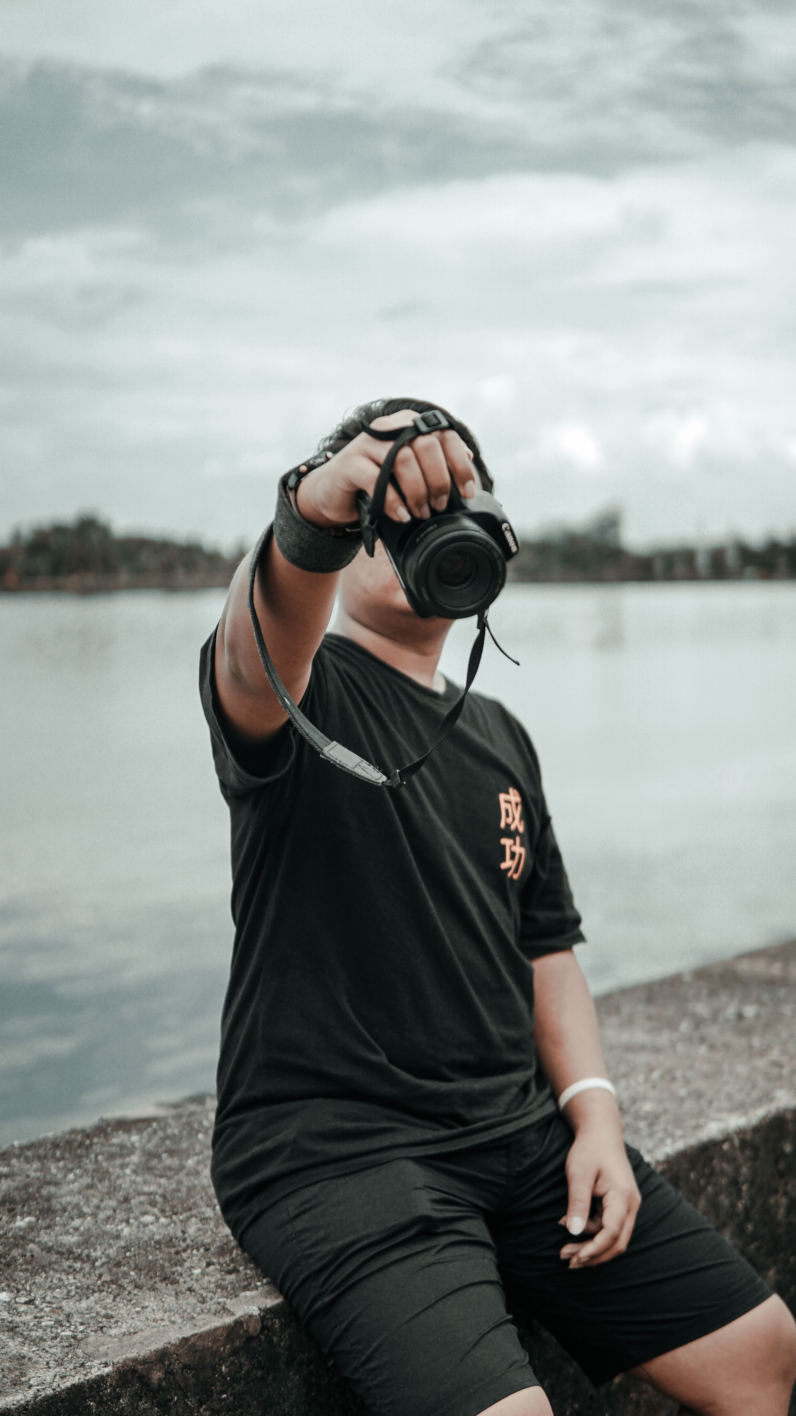 A young man sits by the lake holding a camera against a cloudy sky.