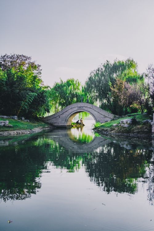 Free Serene view of a classical stone bridge reflecting on a calm river surrounded by lush greenery. Stock Photo