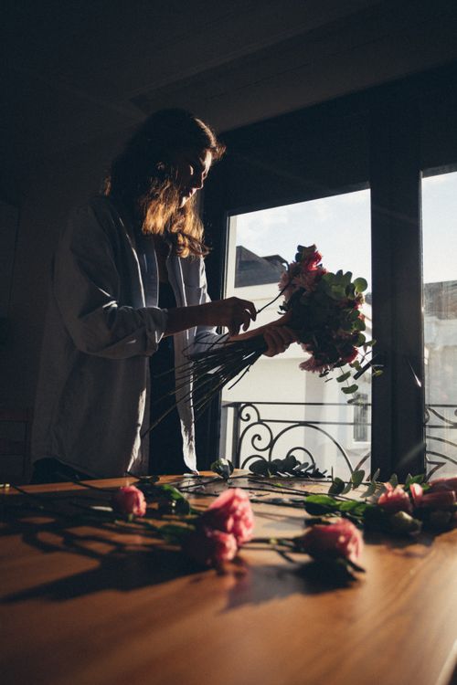 Free A woman arranges a bouquet of flowers on a table lit by warm sunlight, creating a calm and artistic atmosphere. Stock Photo