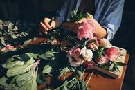 A florist arranging a beautiful bouquet of pink and white flowers using green leaves on a wooden table.
