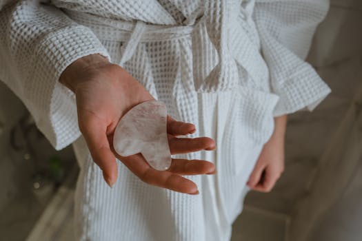 Close-up of a woman's hand holding a gua sha stone, wearing a white bathrobe for skincare routine.