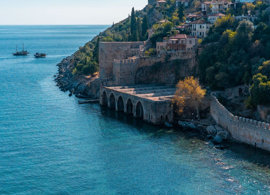 Weathered stone fortress located on rocky seashore with green trees and small settlement with residential buildings near calm sea with boats