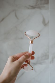Close-up of a hand holding a rose quartz facial roller against a marble background.