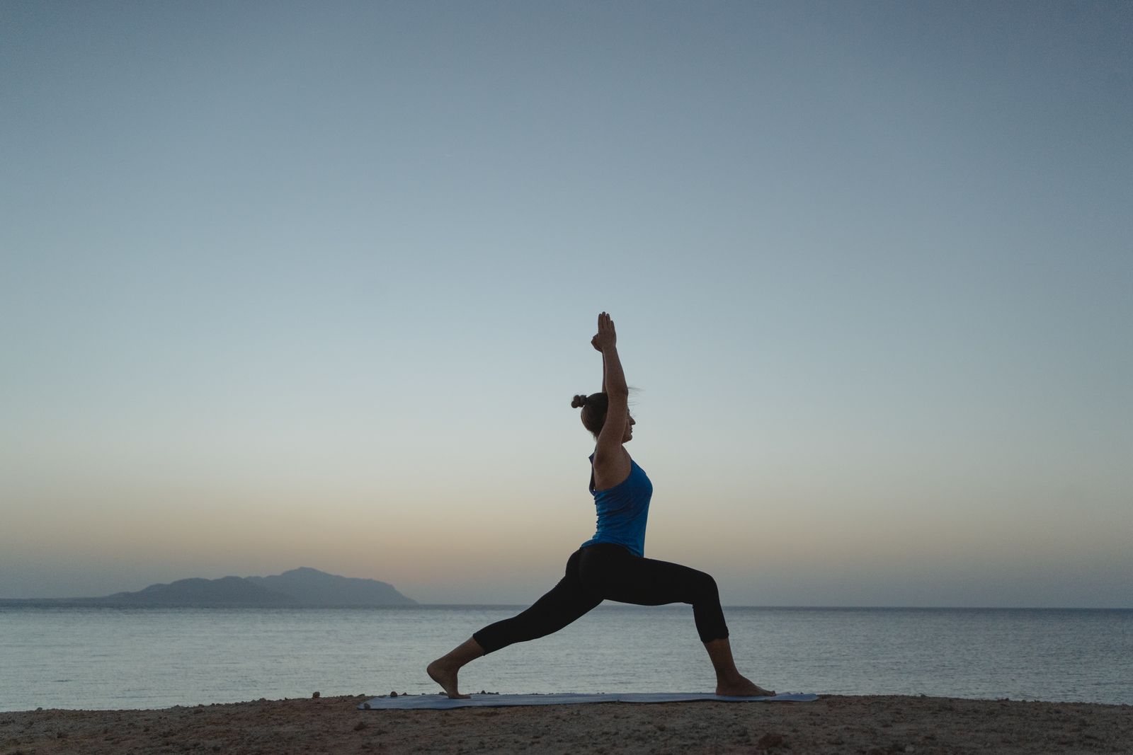 Woman practicing Warrior pose in morning yoga on a beach mat at sunrise with calm ocean water in the background