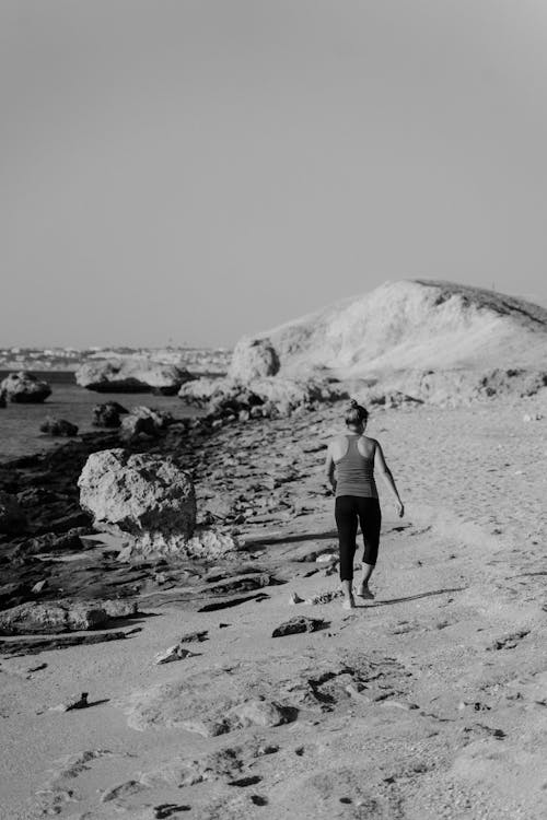 Grayscale Photo of Woman in Black Tank Top and Shorts Walking on Sand
