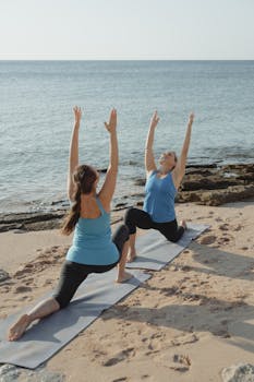 Two women performing yoga Warrior I pose on a serene beach, promoting fitness and relaxation.
