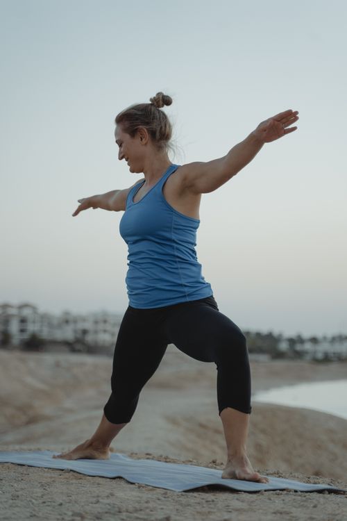 Free Caucasian woman performing yoga in a warrior pose on a sandy beach during the tranquil evening. Stock Photo