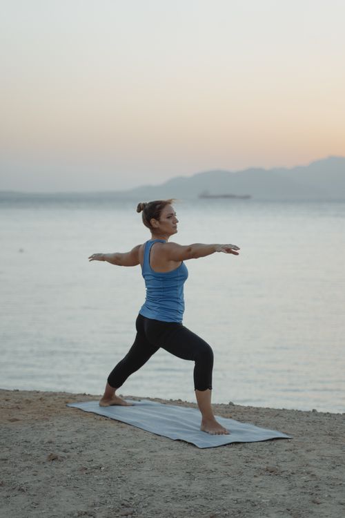 Free Woman performing Warrior 2 pose on the beach during twilight, combining fitness and tranquility. Stock Photo