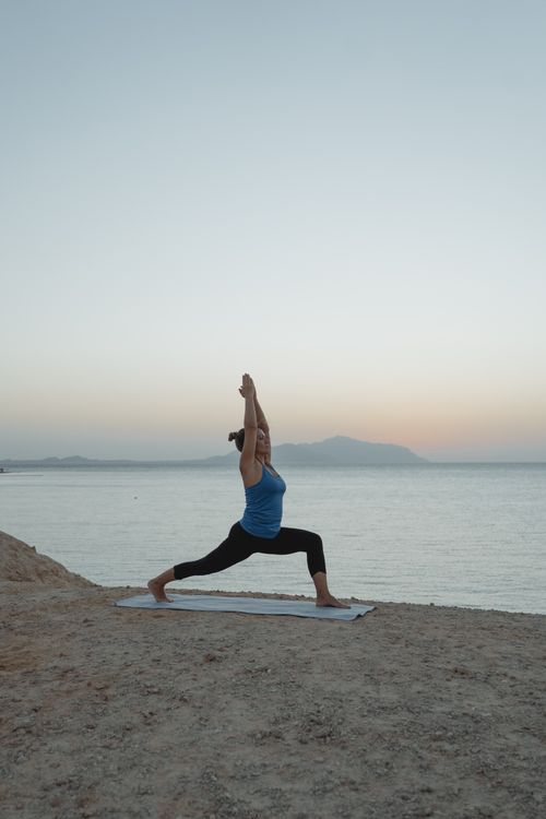 Free A woman performs Warrior 1 yoga pose on a yoga mat at twilight by the sea, promoting tranquility and fitness. Stock Photo