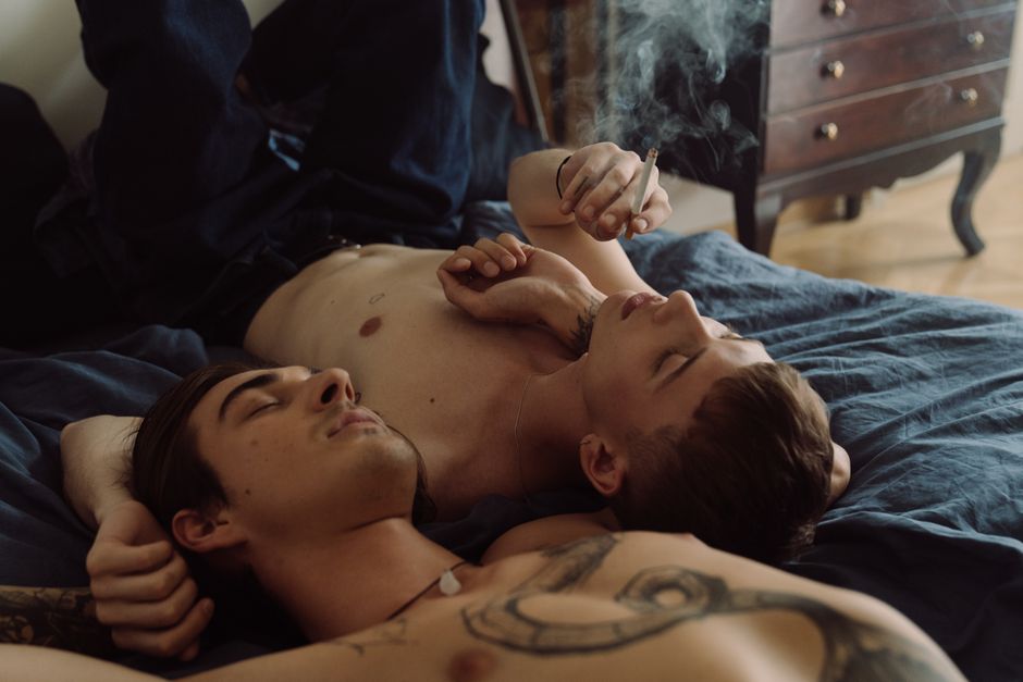 Two shirtless men lying together in a bedroom, one smoking, creating an intimate and relaxed atmosphere.