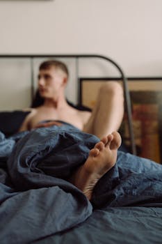 Shirtless man relaxes in bed under a blue blanket, capturing a calm morning vibe.