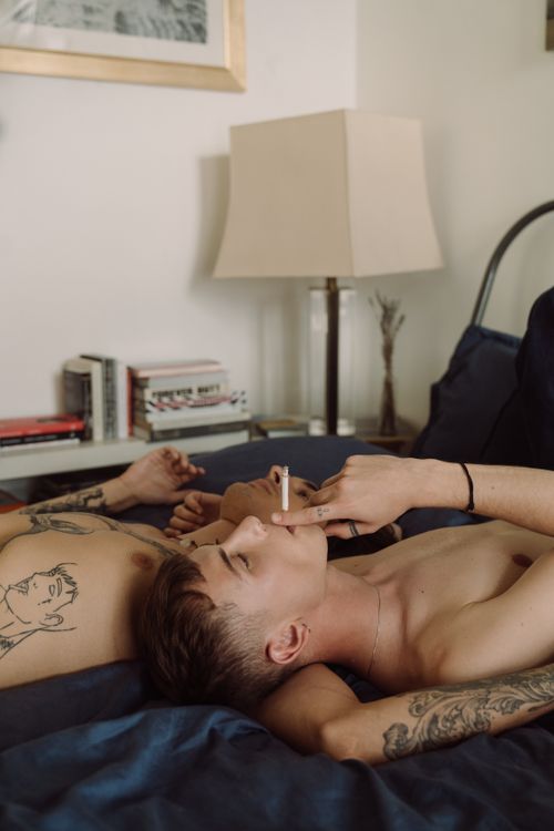 Free An intimate and relaxed scene of a tattooed couple lounging shirtless in a modern bedroom, conveying love and connection. Stock Photo
