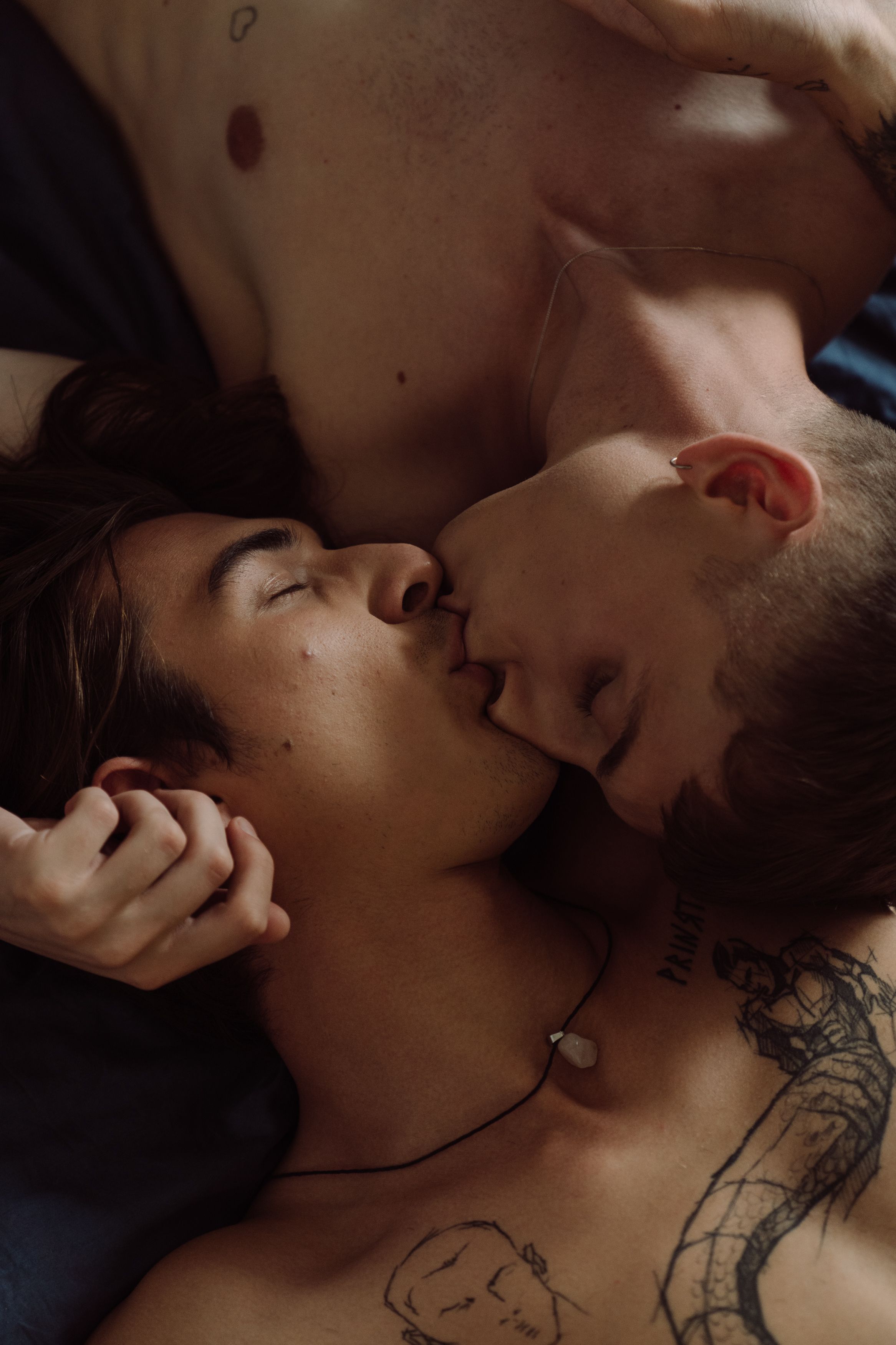 A tender, intimate kiss between two shirtless men lying closely together.
