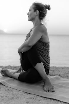 Woman practicing yoga pose on a mat by the seaside at sunset in black and white.