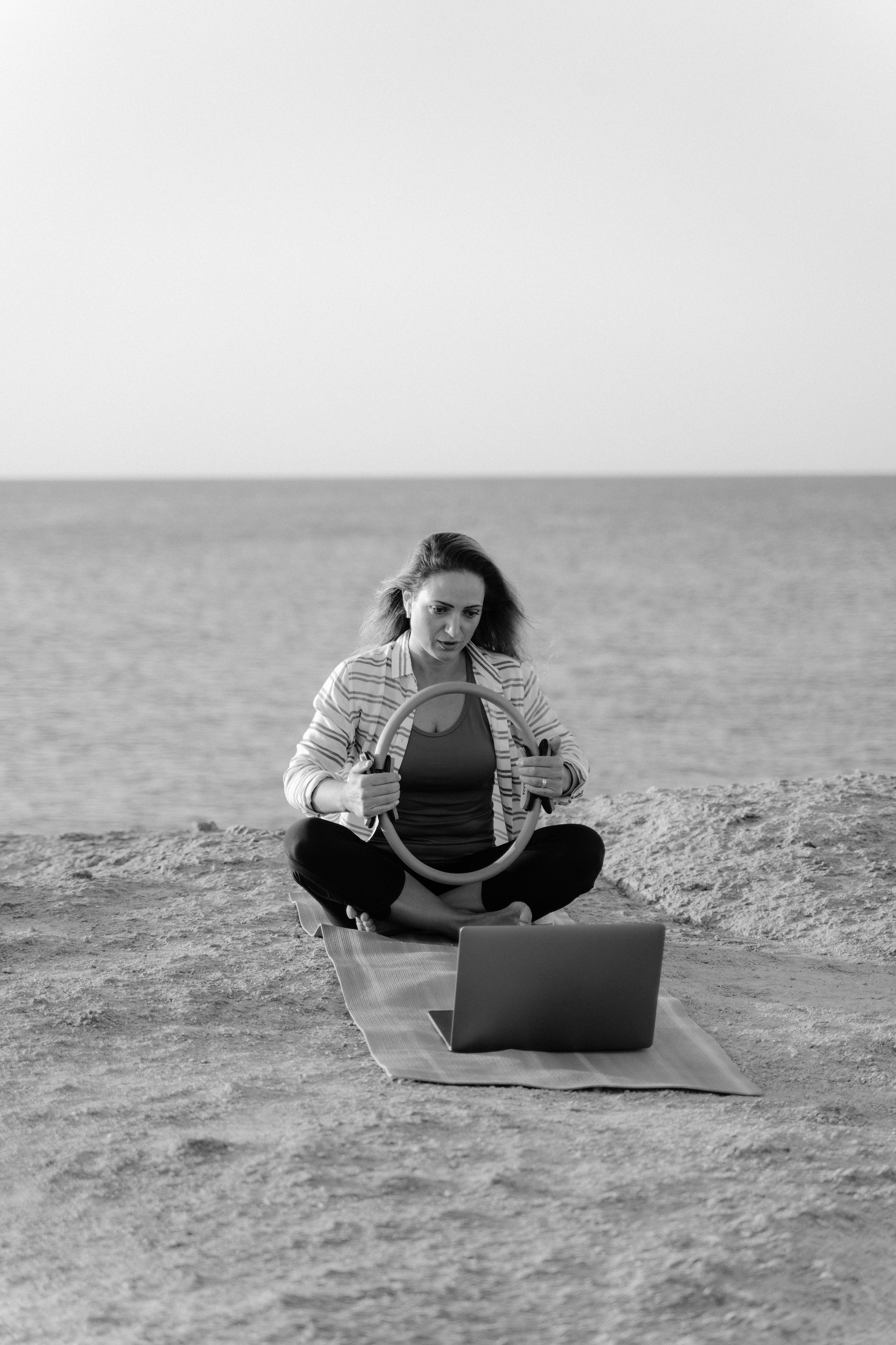 Monochrome photo of woman doing yoga with a ring and laptop by the sea.