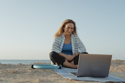 Woman in White and Blue Striped Long Sleeve Shirt Sitting on Yoga Mat In Front of Laptop 