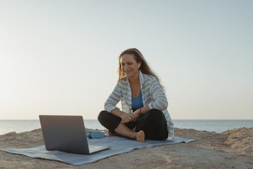 Free Woman in White Long Sleeve Shirt and Black Pants Sitting on Gray Sand Using Macbook Stock Photo