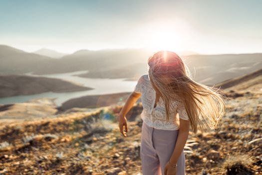 A blonde woman stands on a hilltop enjoying a stunning sunset over rolling hills and water, embracing nature.