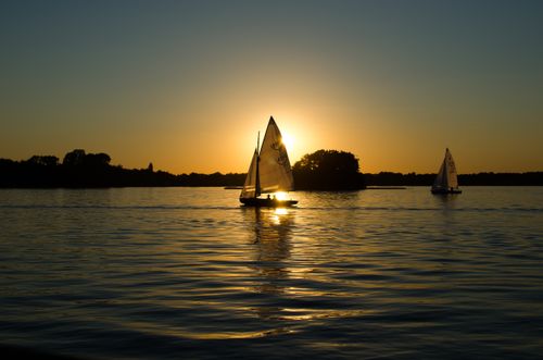 Free Silhouetted sailboats glide peacefully over the water beneath a vibrant sunset, casting serene reflections. Stock Photo