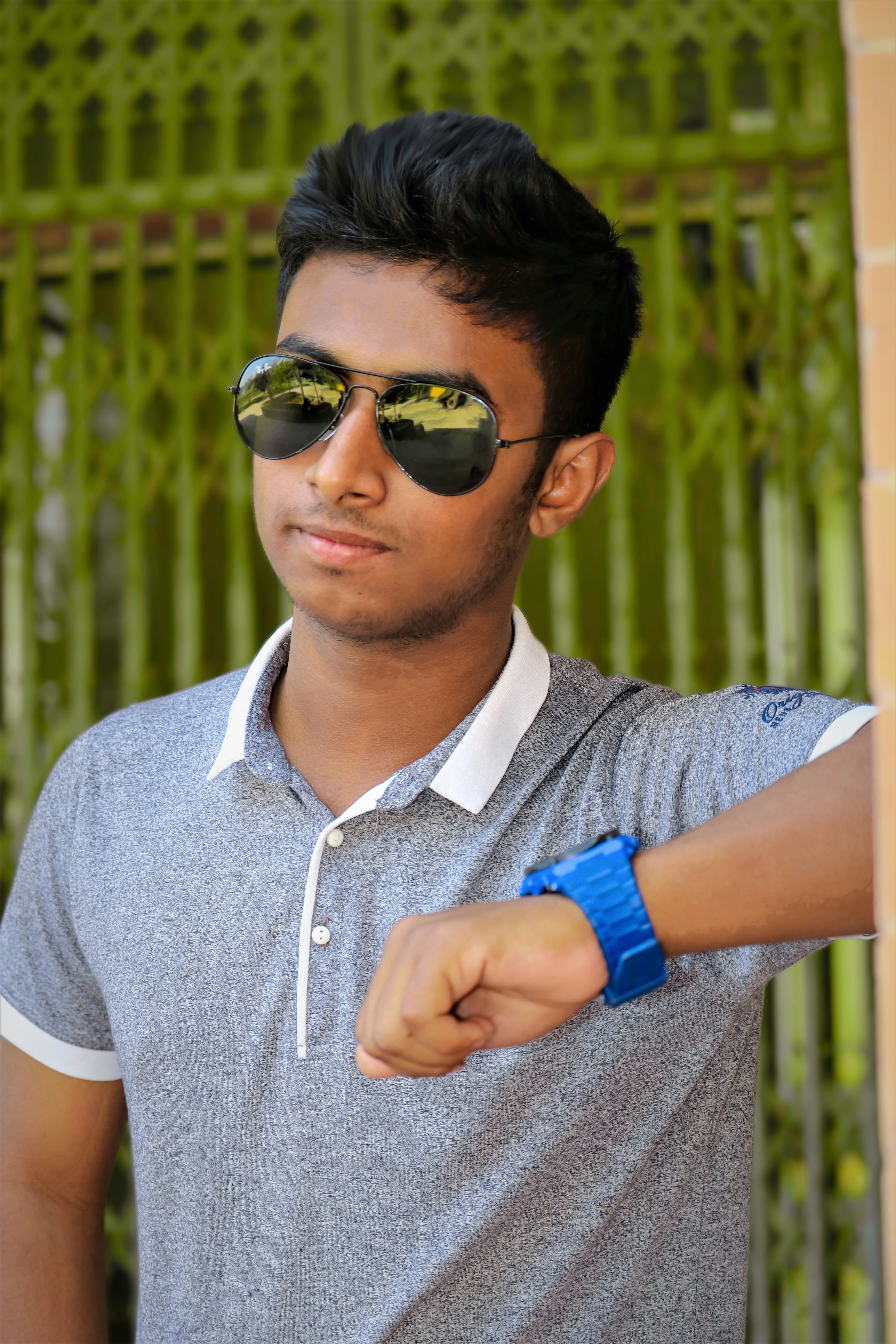A young man leans casually against a wall, wearing sunglasses and a wristwatch.