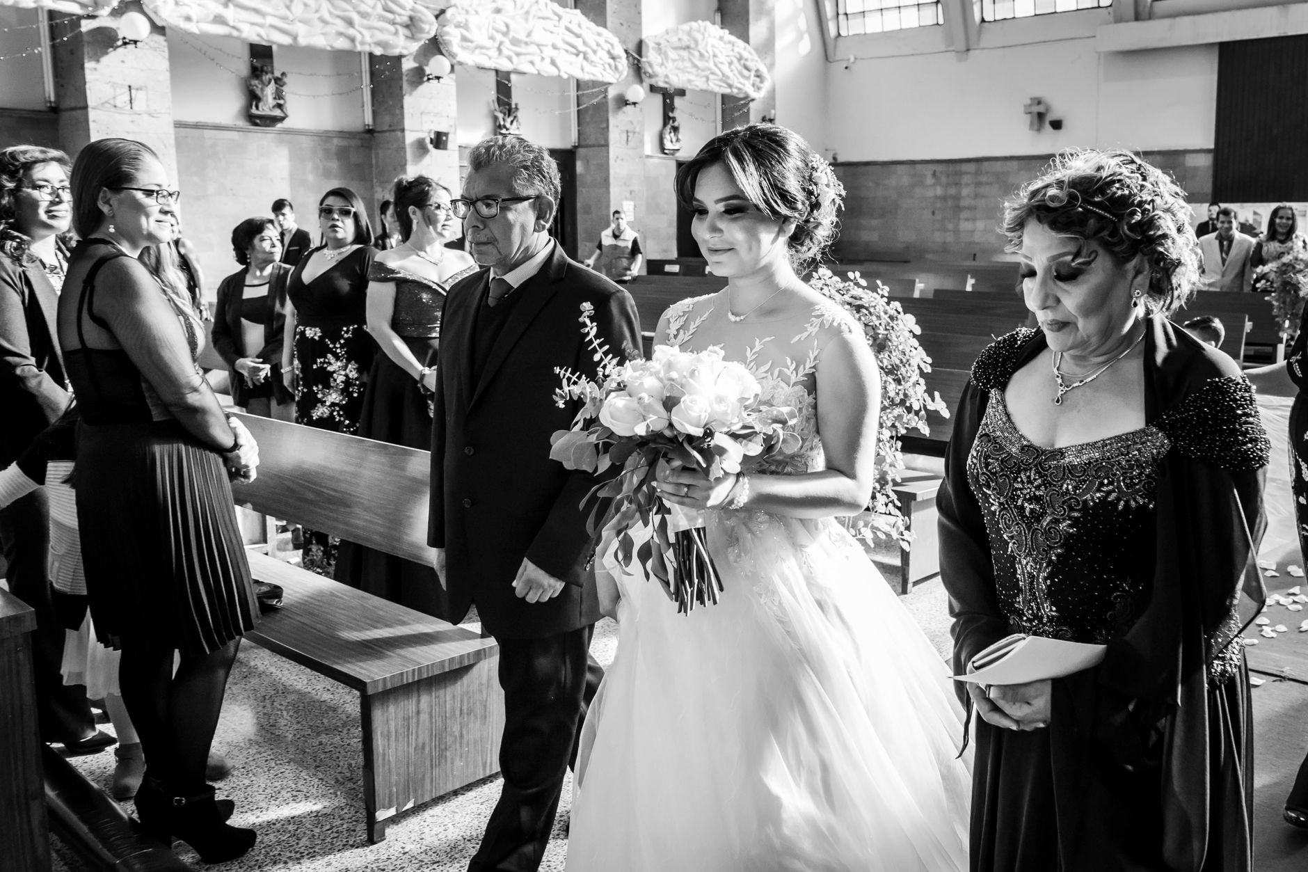 Emotional black and white photo of a bride walking down the aisle with her parents in a church wedding ceremony.
