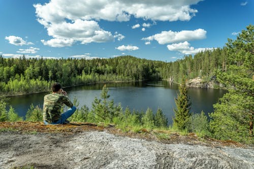 Free Man in Green Camouflage Long Sleeve Shirt Sitting on Rock Near Lake Stock Photo