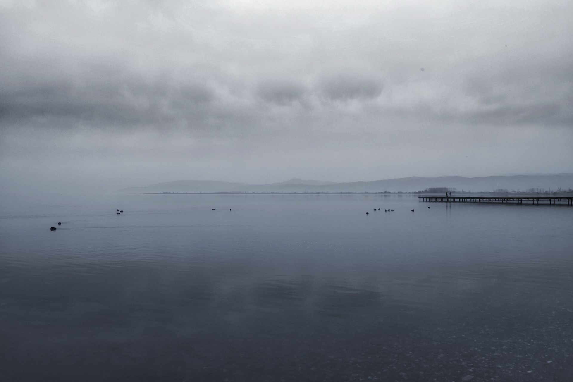 Tranquil overcast view of İznik lake with a distant dock under cloudy skies.