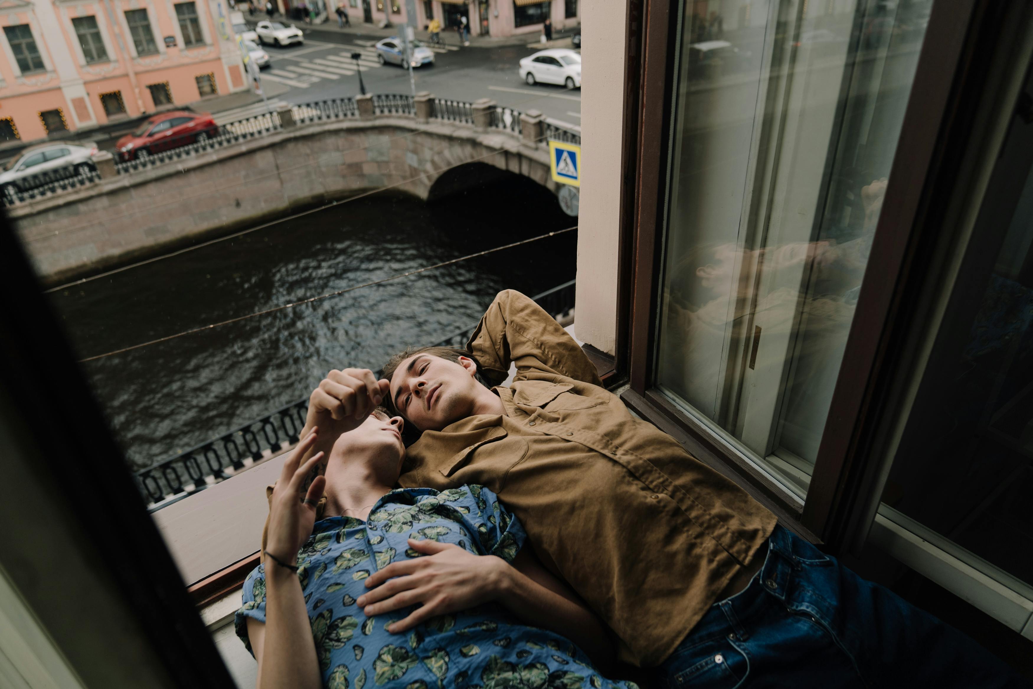 A loving couple embraces on a windowsill overlooking a river in a city.