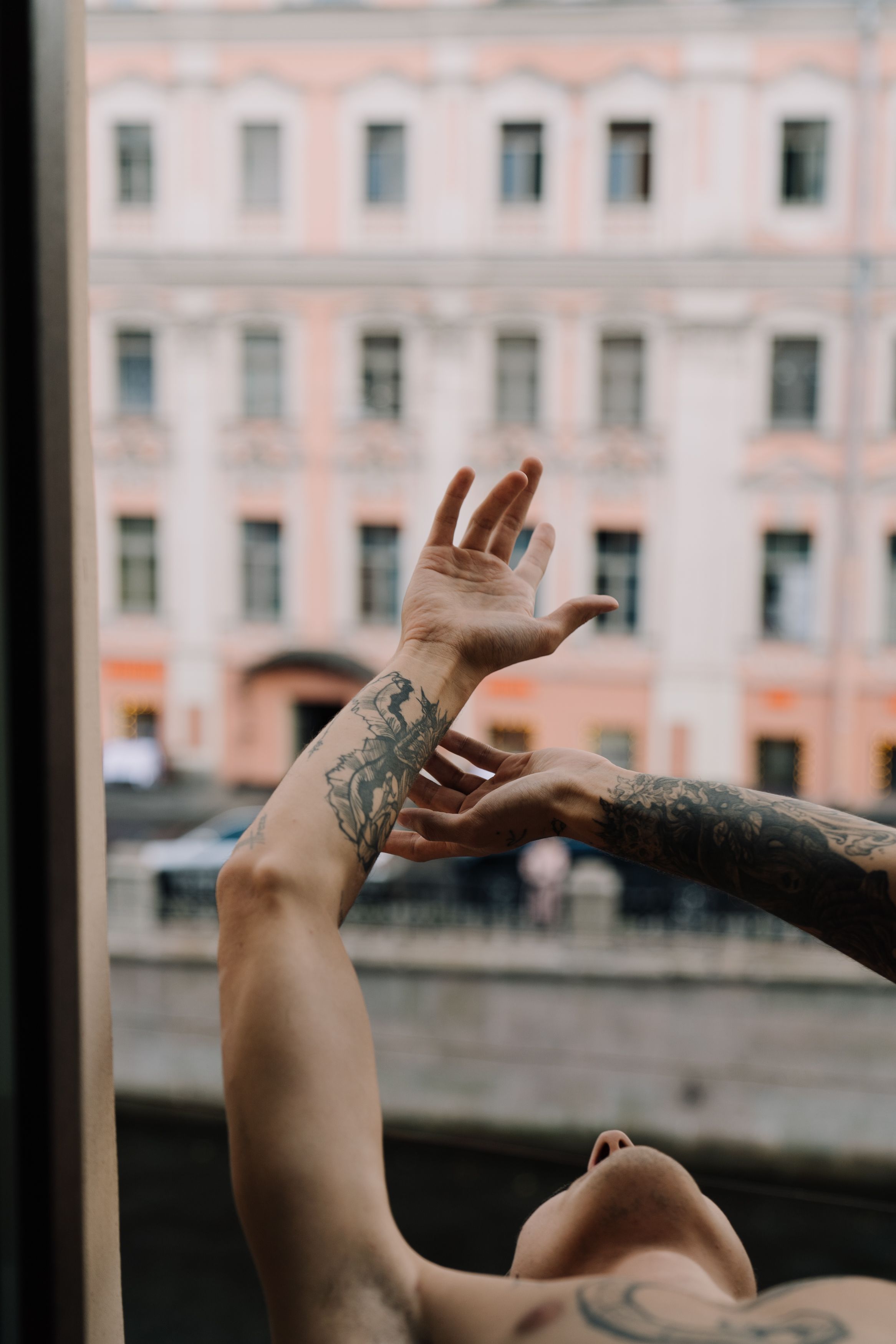 A person reaches out from a window, showcasing tattooed arms against a classic facade.