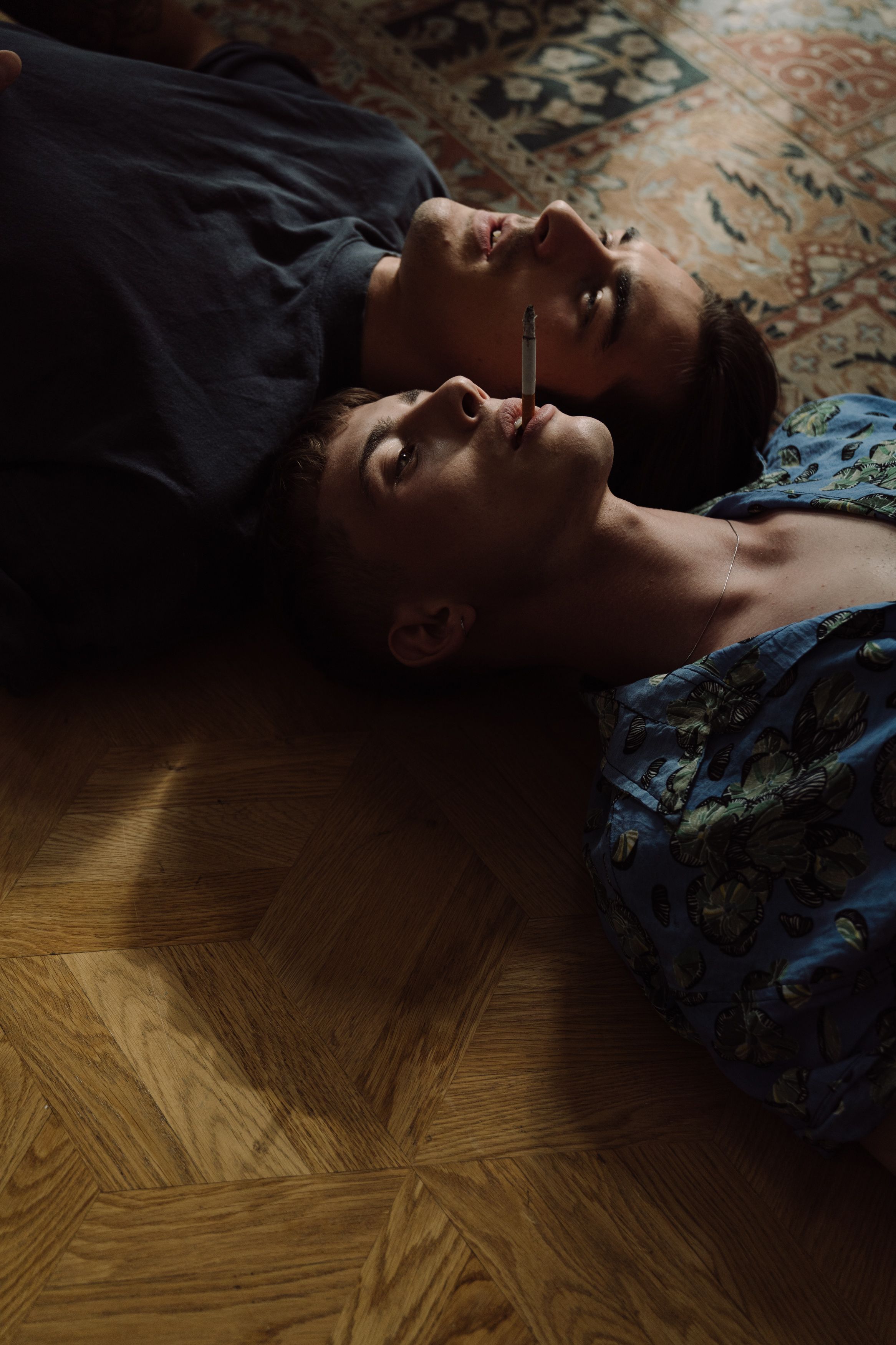A close-up of two men lying together on a wooden floor, sharing a cigarette in an intimate setting.
