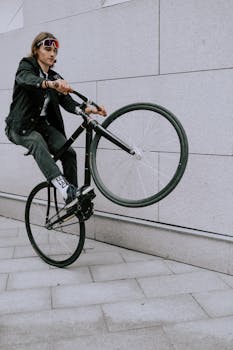 Man in black outfit performing a wheelie on a bicycle against a grey wall.