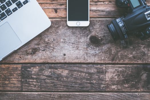 Flat lay of rustic wooden desk with laptop, smartphone, and camera.