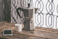 木桌上放着摩卡壶和咖啡杯，还有一个智能手机，非常适合舒适的早晨