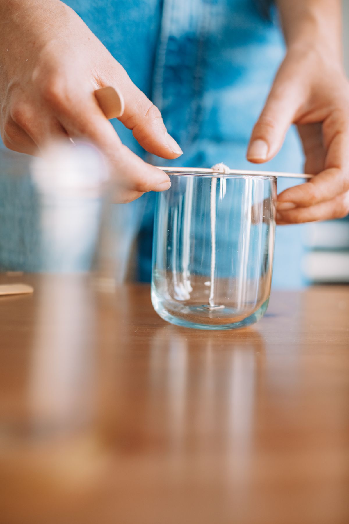 Hands preparing a candle wick inside a glass jar on a wooden table