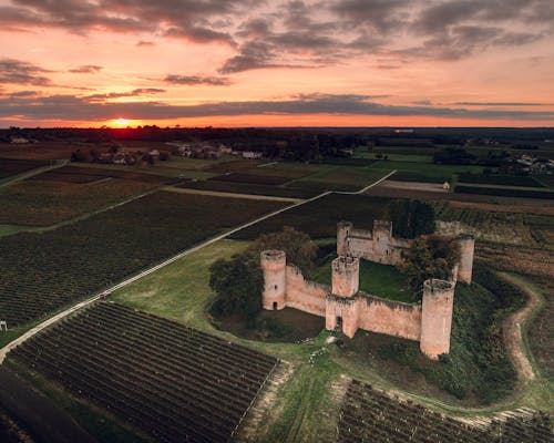 Drone view of ancient stone temple surrounded by black plowed gardens under colorful sky at sunset