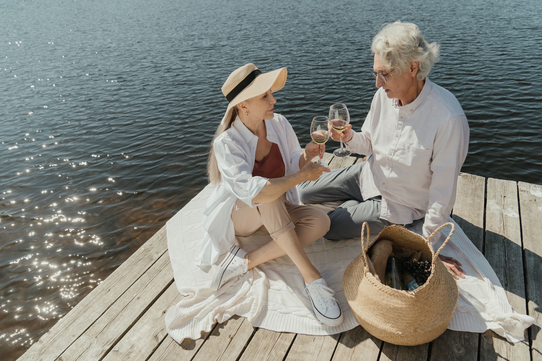 An elderly couple toasting wine glasses on a sunny lakeside pier, enjoying a picnic.