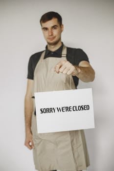 Photo by Gustavo Fring A man wearing an apron holds a 'Sorry We're Closed' sign, indicating shop closure.