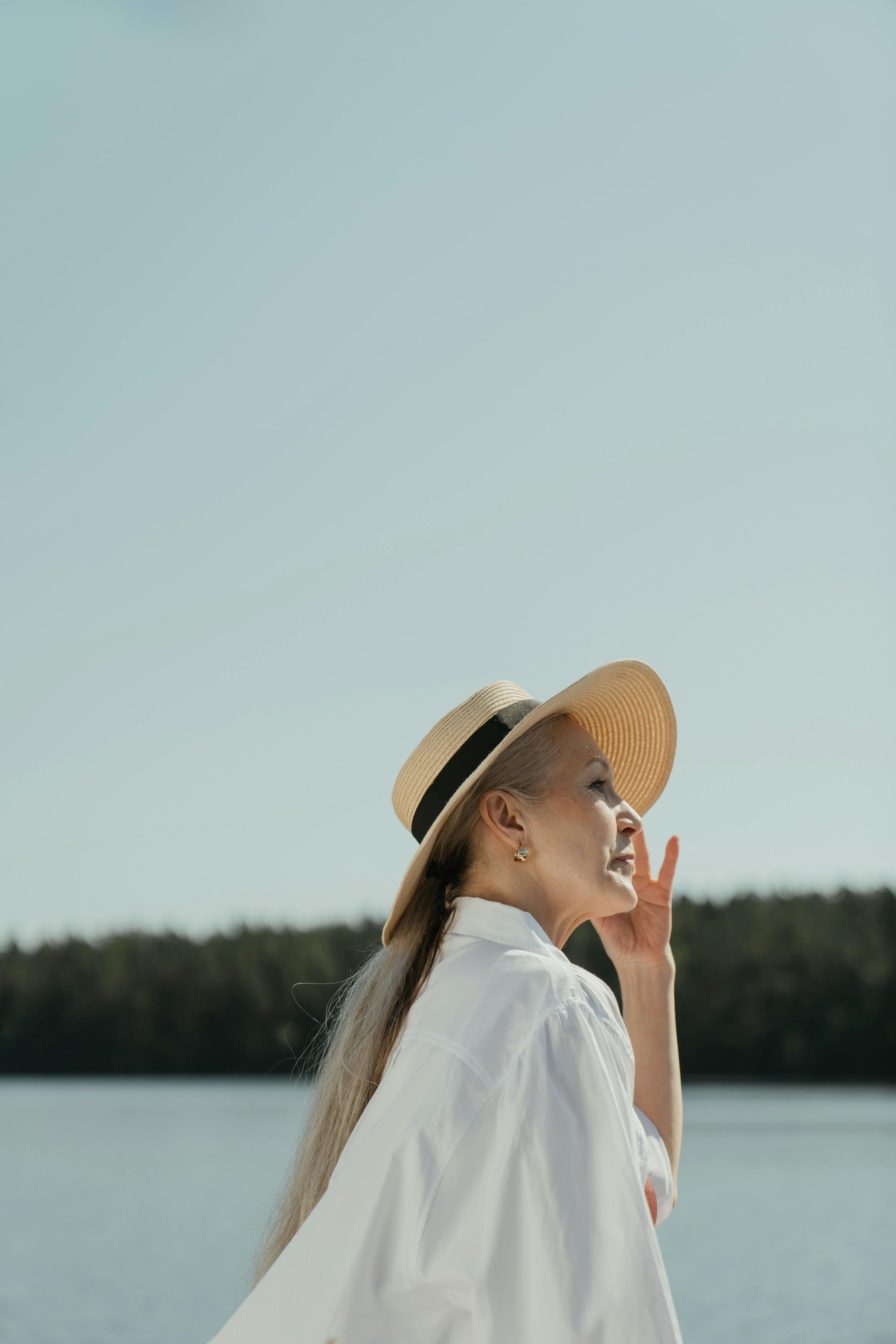 在晴朗的天空下，一位戴着太阳帽的时尚老妇人侧影，正欣赏着湖边宁静的景色