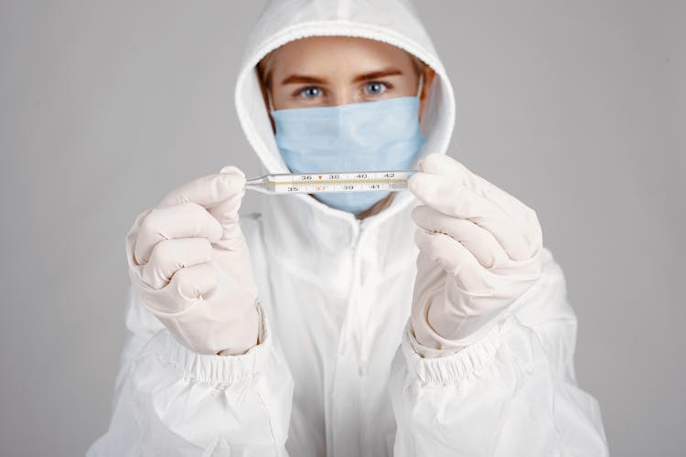 Person In Face Mask And Protective Suit Holding Thermometer