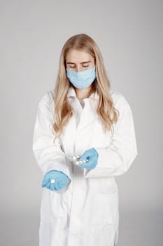 Portrait of a female doctor in a white coat and blue mask holding pills, in a studio setting.