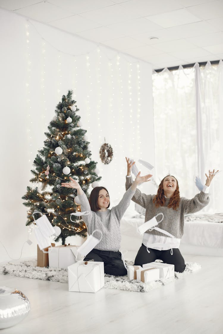 Women Sitting On Floor Near Christmas Tree Throwing Face Masks In The Air