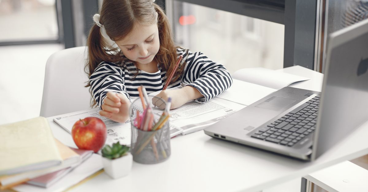 A young girl studies at home using a laptop, engaged with her homework and school supplies.