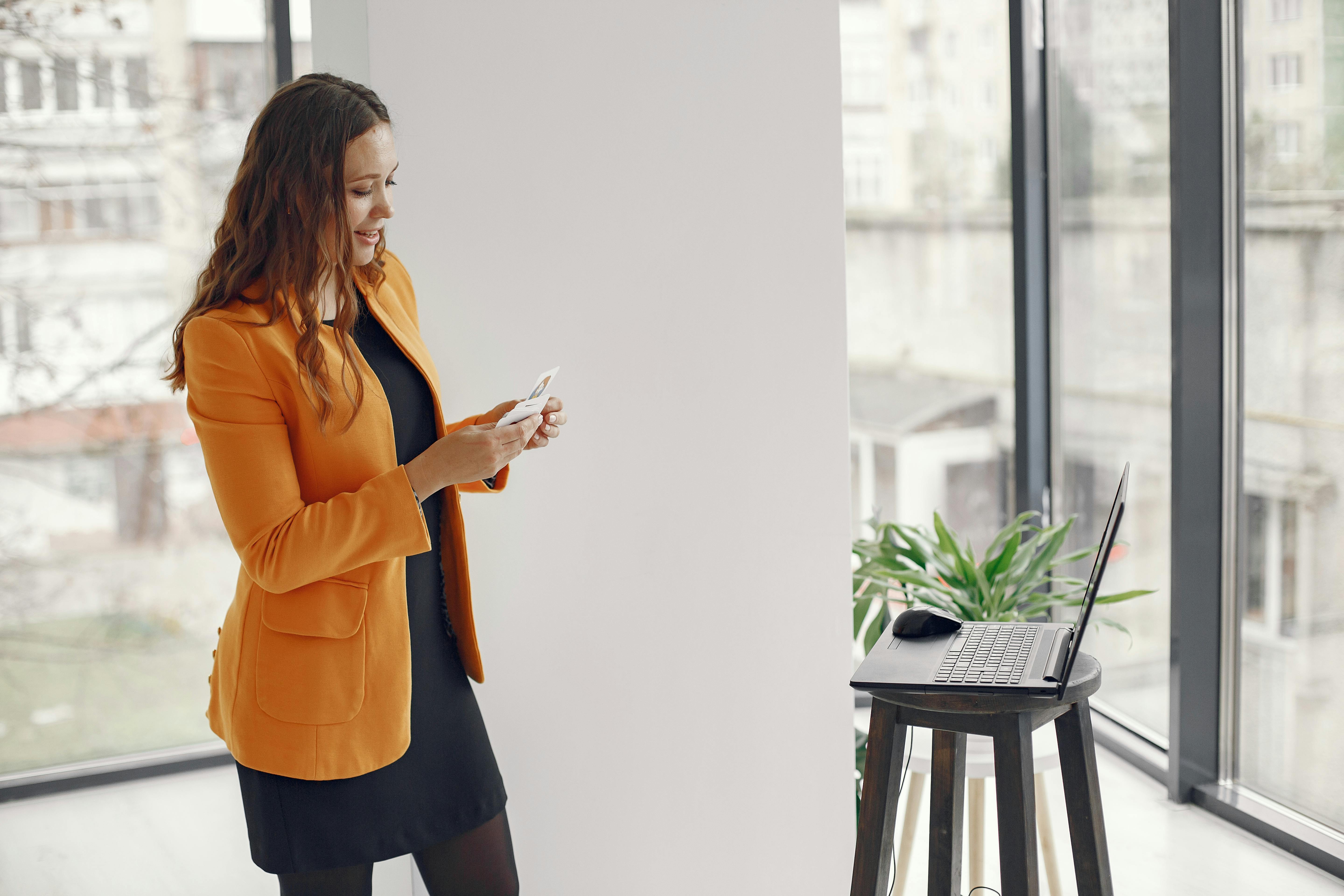 Businesswoman with orange jacket holds cards and stands by laptop in modern office.