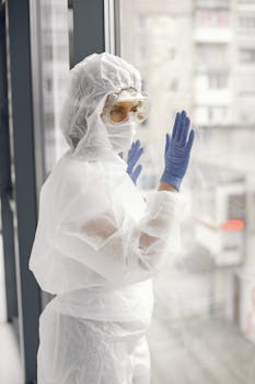 Healthcare professional in protective gear stands by a window, symbolizing safety and care.