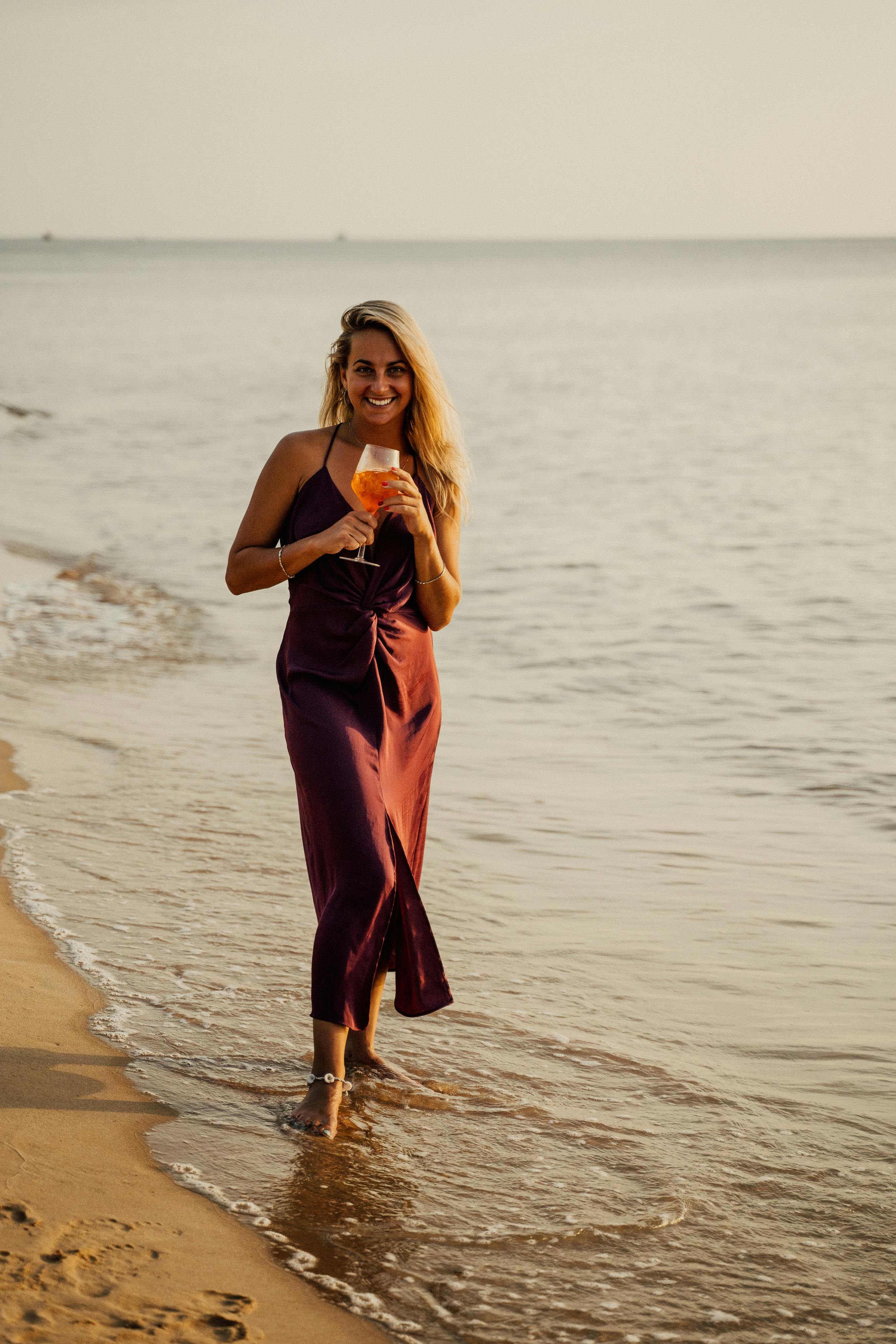 a woman in brown dress holding a glass of drink on the beach