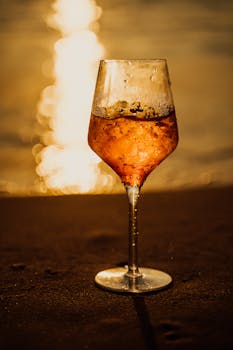 A wine glass filled with a vibrant orange drink on a sandy beach during sunset.