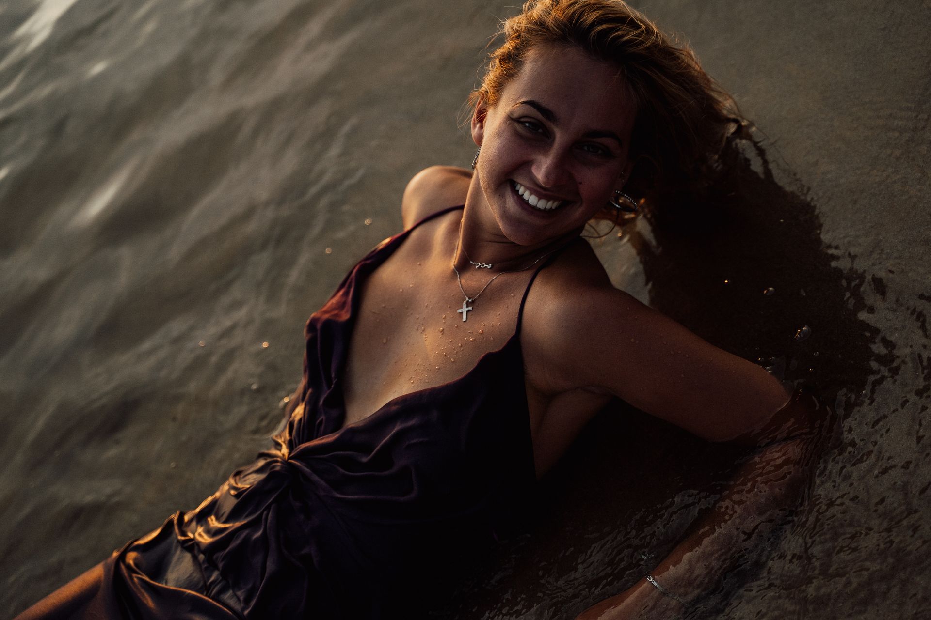 A happy woman in a dress relaxes in shallow beach waters, smiling under warm sunset light.