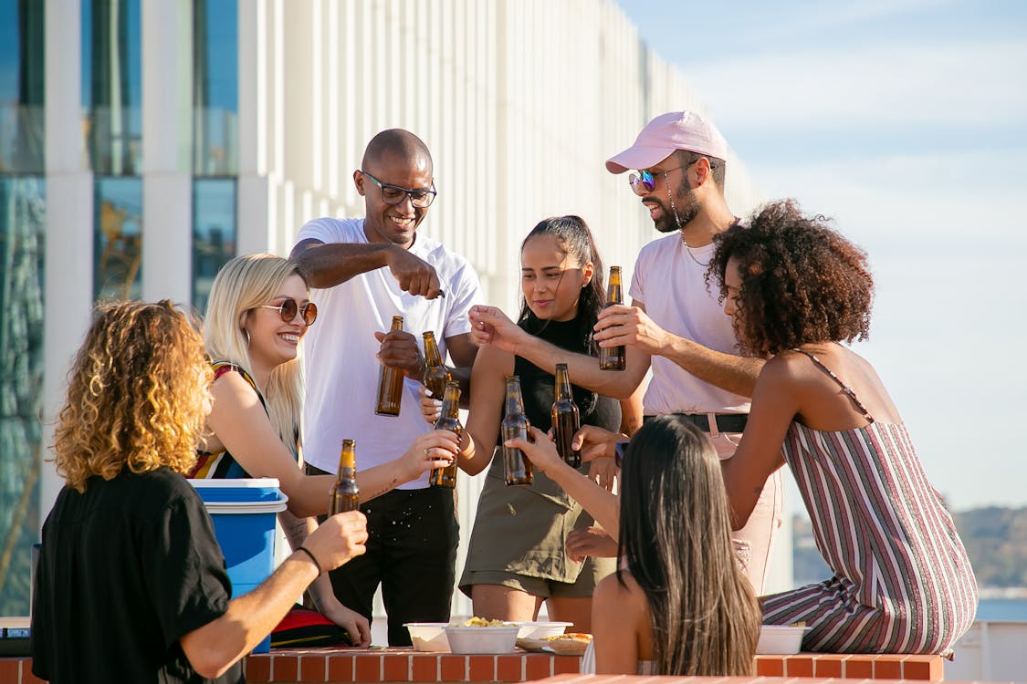 Free Group of cheerful multiracial friends in casual summer outfits toasting with beer bottles while having party on rooftop in urban environment Stock Photo