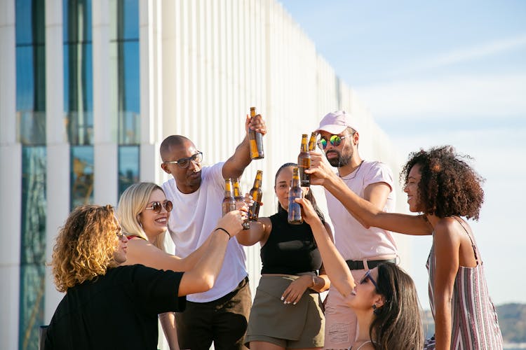 Joyful Diverse Friends Clinking Beer Bottles On Rooftop